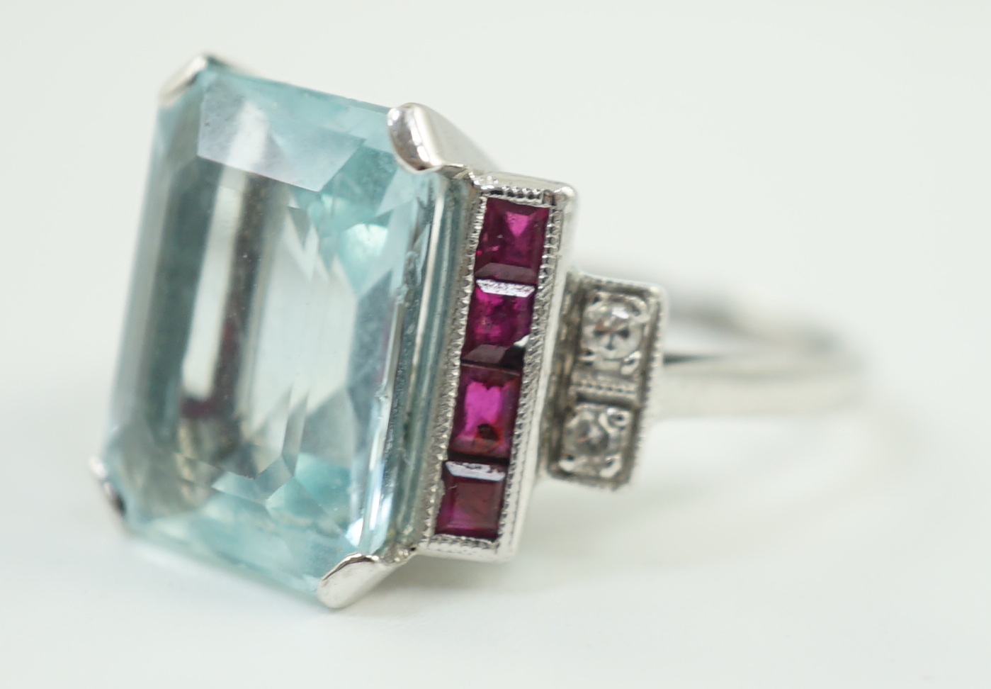 A platinum and single stone emerald cut aquamarine set dress ring, with ruby and diamond set stepped shoulders